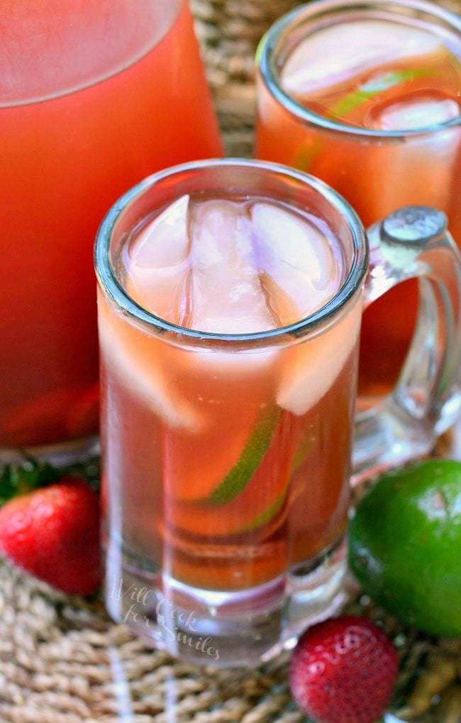 Top view of Strawberry Lime Infused Iced Tea in a glass mug. There is floating ice, as well as strawberry and lime slices in the glass. An additional mug and pitcher are in the background. A couple strawberries and lime lay on the table.