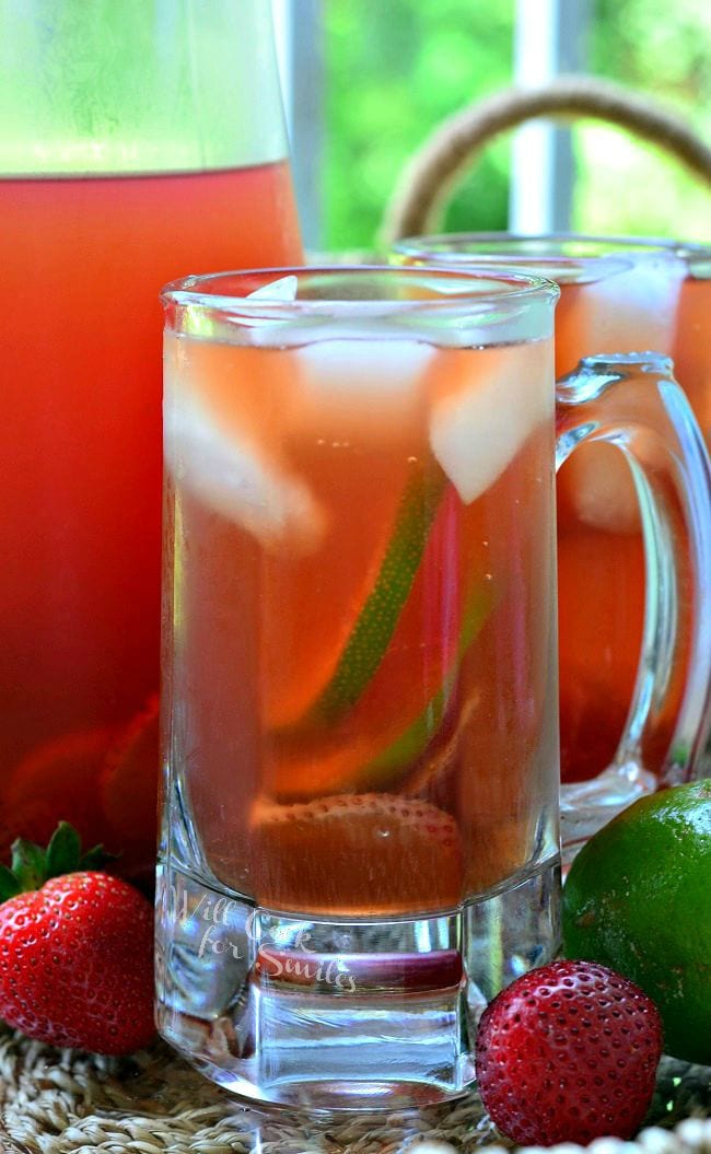 Strawberry Lime Infused Iced Tea is presented in a glass mug. There is floating ice, as well as strawberry and lime slices in the glass. An additional mug and pitcher are in the background. A couple strawberries and lime lay on the table.