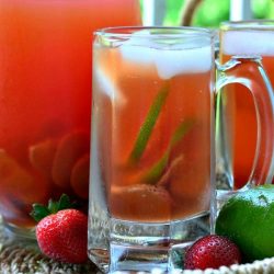 Clear glass beer mug filled with strawberry lime infused iced tea in front of another mug and pitcher filled with tea viewed close up