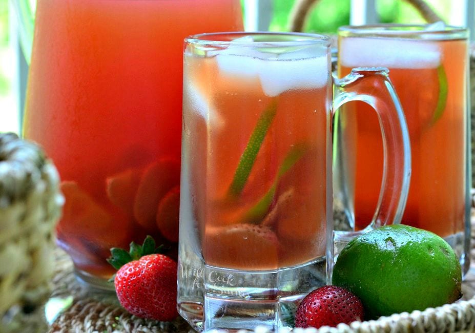 A side view of the Strawberry Lime Infused Iced Tea in a glass mug. There is floating ice, as well as strawberry and lime slices in the glass. An additional mug and pitcher are in the background. A couple strawberries and lime lay on the table.