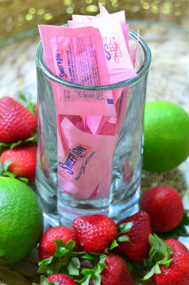 A glass mug is filled with Sweet N'Low packets. The mug is surrounded by strawberries and limes.