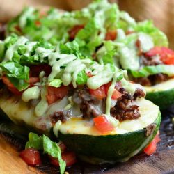 2 taco stuffed zucchini boats on a wooden board topped with avocado dressing viewed close up
