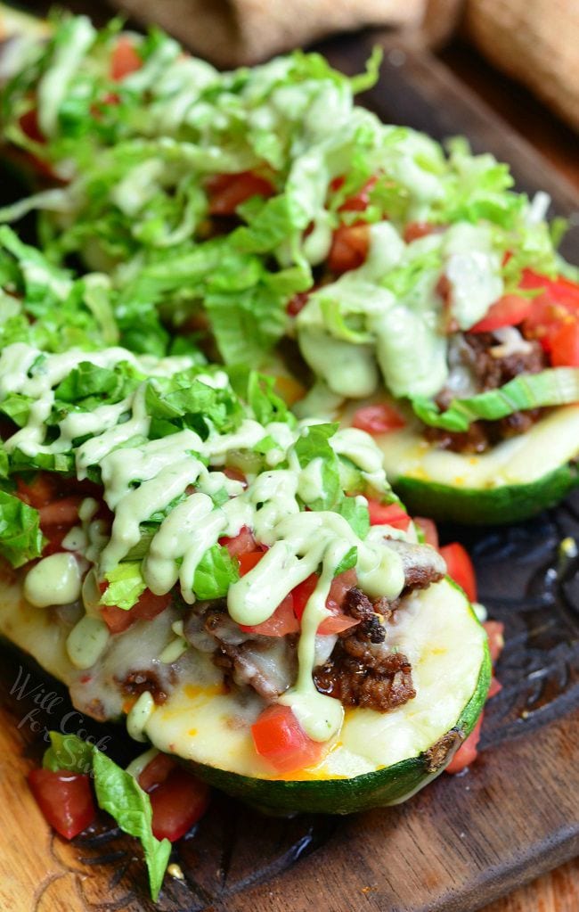 More of a top view of two Taco Stuffed Zucchini Boats on a wooden board. They are topped with lettuce, tomatoes, and drizzled with avocado dressing.