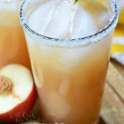 2 pint glasses filled with white peach margarita on a wooden serving tray wtih slice peaches in foreground