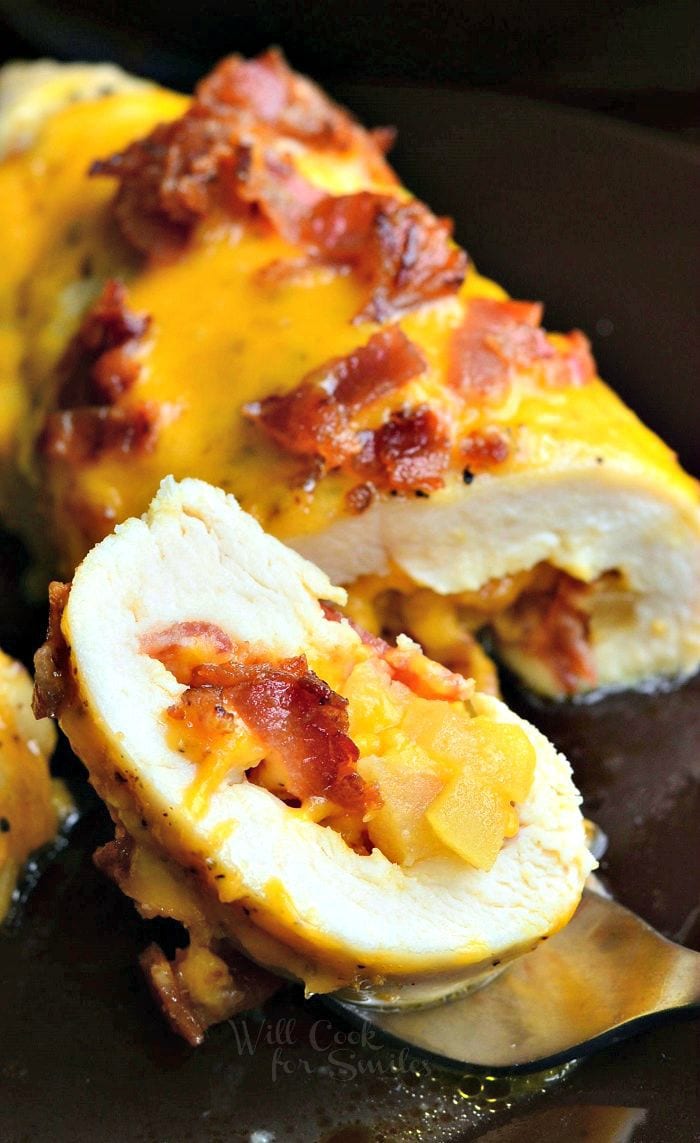 On a fork, there's a slice of chicken breast that's been stuffed with apples, bacon, and cheese. Cheese and bacon appear on top of the remaining chicken breast in the background.