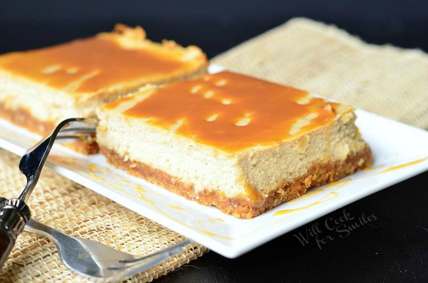 Caramel Macchiato Cheesecake Bars are on a white plate. Caramel is drizzled on top of the bars, as well as on the plate. Forks are criss-crossed to the left side of the plate.