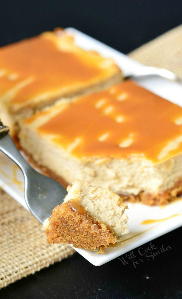 Caramel Macchiato Cheesecake Bars are on a white plate. Caramel is drizzled on top of the bars, as well as on the plate. A fork holds a bite of one of the bars.