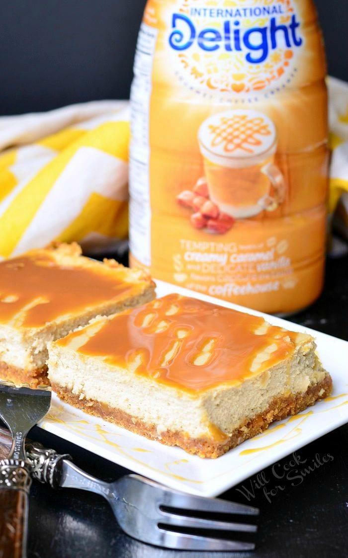 Caramel Macchiato Cheesecake Bars are on a white plate with the forks set to the side. Caramel is drizzled on top of the bars, as well as on the plate. Caramel coffee creamer is sitting on the table in the background.