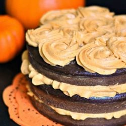 close up view of chocolate cake with pumpkin cream cheese frosting on orange doilies with 2 pumpkins in the background