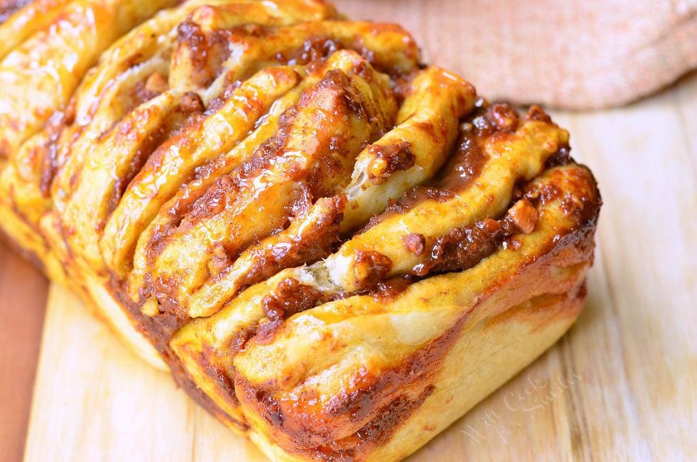 Easy Caramel Pumpkin Pull Apart Bread sits on a cutting board. Each layer is slathered in pumpkin, caramel, and pecans. There is a caramel sauce drizzled on top as well.