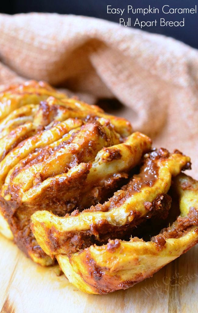 Easy Caramel Pumpkin Pull Apart Bread sits on a cutting board. The first two layers slightly pulled. Each layer is slathered in pumpkin, caramel, and pecans. There is a caramel sauce drizzled on top as well.