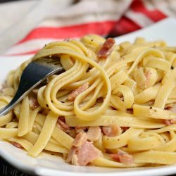 white square plate with fettuccine whit bourbon bacon sauce on a wooden table as a fork leans on the left side of the plate