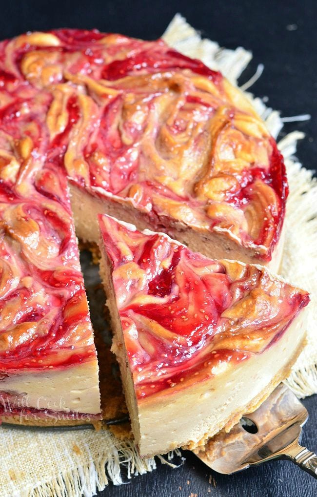 A pie server is removing a large piece of the Peanut Butter and Jelly Cheesecake from the whole. This cheesecake has beautiful swirls of peanut butter and red jelly, with seeds, on top.