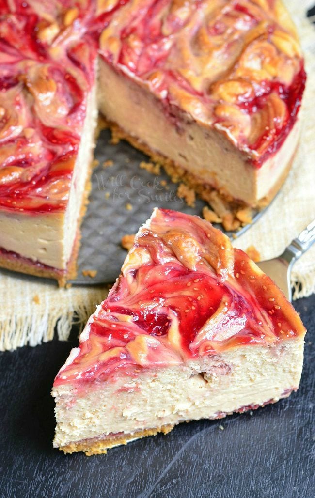 A large piece of the Peanut Butter and Jelly Cheesecake was removed from the whole. This piece sits out-front at an angle. This cheesecake has beautiful swirls of peanut butter and red jelly, with seeds, on top.