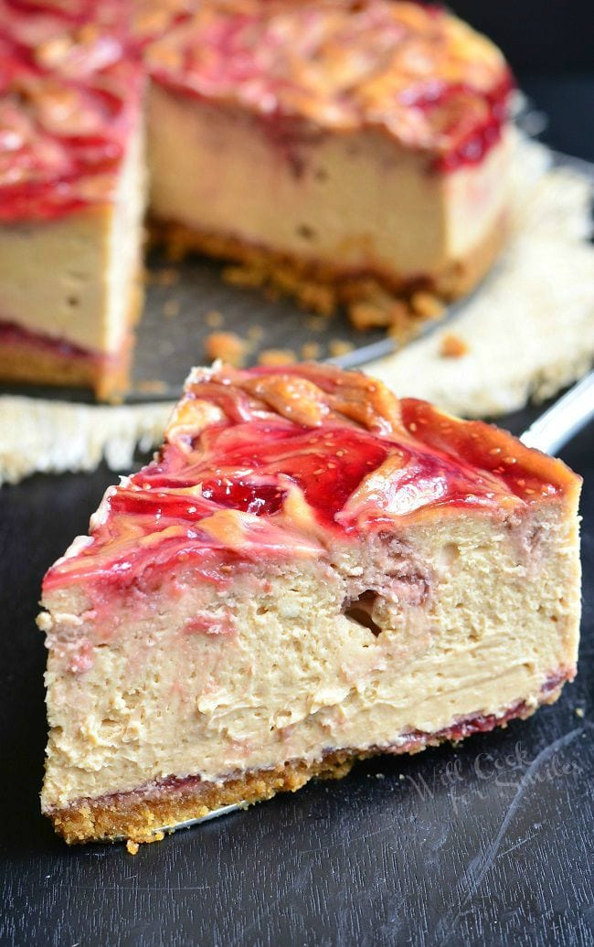 A large piece of the Peanut Butter and Jelly Cheesecake was removed from the whole. This piece sits out-front at an angle. This cheesecake has beautiful swirls of peanut butter and red jelly, with seeds, on top.
