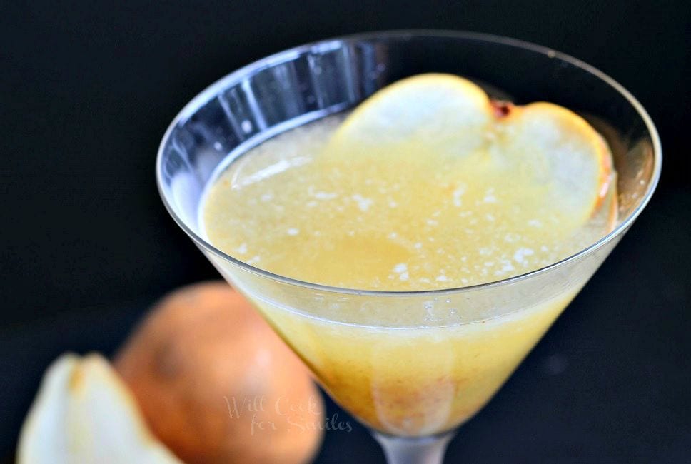 The Pear Martini is in a tall martini glass. The liquid is yellow-ish in color. Pulp has collected towards the bottom of the glass. A thinly sliced piece of pear is on the inside of the glass for garnish.
