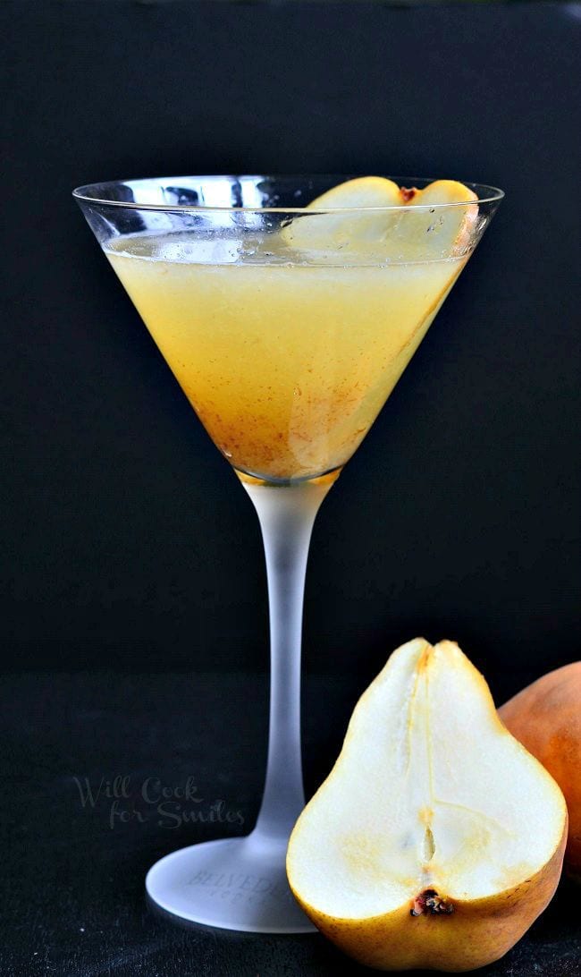 The Pear Martini is in a tall martini glass. The liquid is yellow-ish in color. Pulp has collected towards the bottom of the glass. A thinly sliced piece of pear is on the inside of the glass and a pear is half sitting next to the glass.