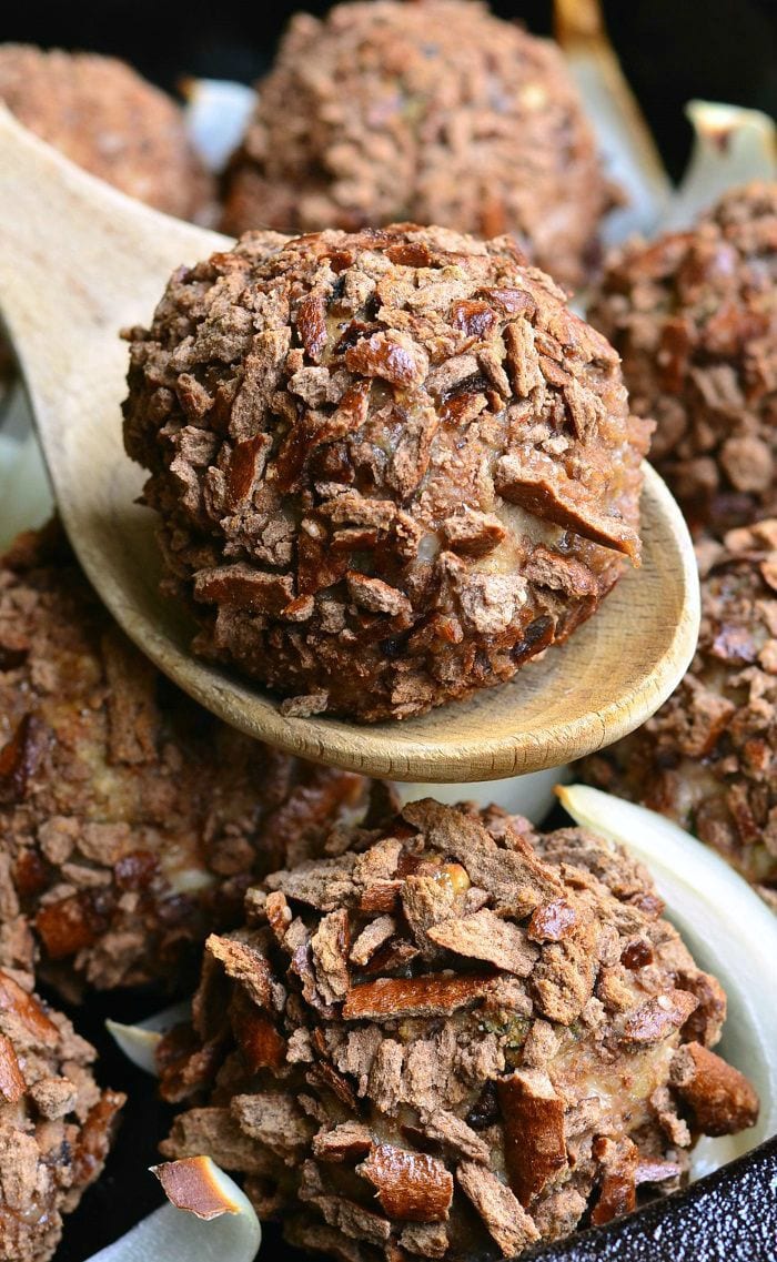 Pretzel Crusted Parmesan Turkey Meatball placed on a wooden spoon. Other Pretzel Crusted Parmesan Turkey Meatballs are below with slices of onion in a black skillet.