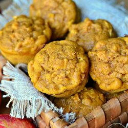 wooden basket filled with pumpkin apple muffins on a wooden table with red apples placed around the basket viewed close up