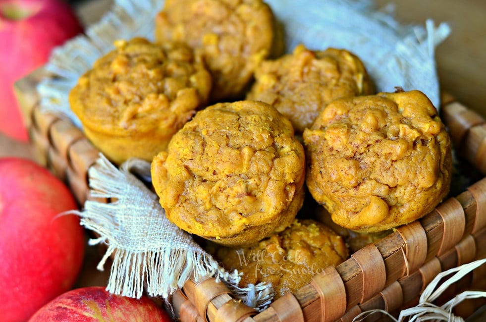wooden basket filled with pumpkin apple muffins on a wooden table with red apples placed around the basket viewed close up