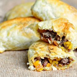 3 pumpkin beef onion hand pies sitting on a tan placemat with the first hand pie broken in half to show inside viewed close up