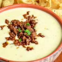 close up view of Queso blanco dip with chorizo in an orange clay bowl on a wooden table with tortilla chips spread across table behind bowl