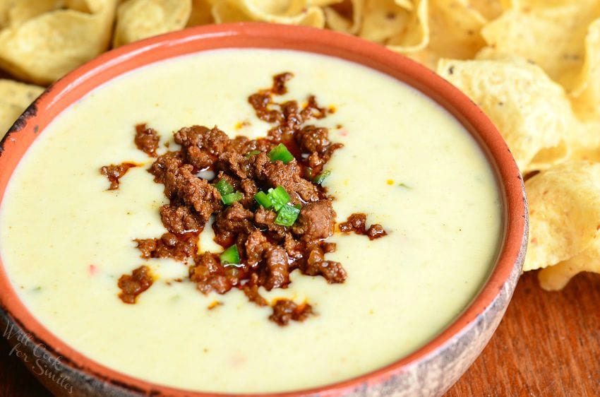 close up view of Queso blanco dip with chorizo in an orange clay bowl on a wooden table with tortilla chips spread across table behind bowl