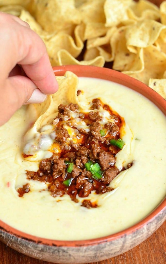 Queso blanco dip with chorizo in an orange clay bowl on a wooden table with tortilla chips spread across table behind bowl as a hand dip a chip in the queso