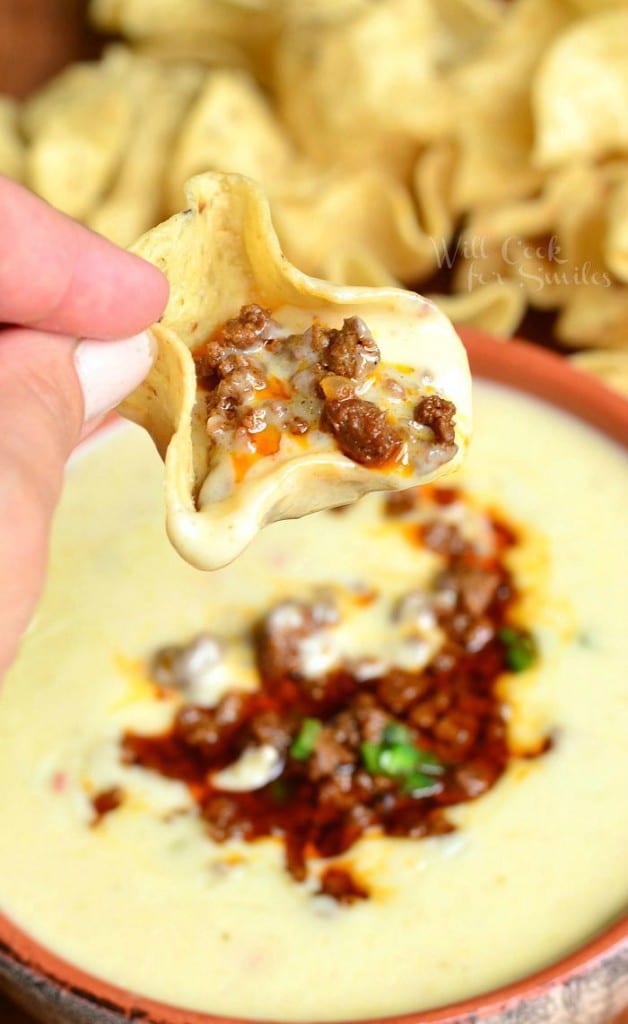 A hand holding 1 chip filled with dip above Queso blanco dip with chorizo in an orange clay bowl on a wooden table with tortilla chips spread across table behind bowl