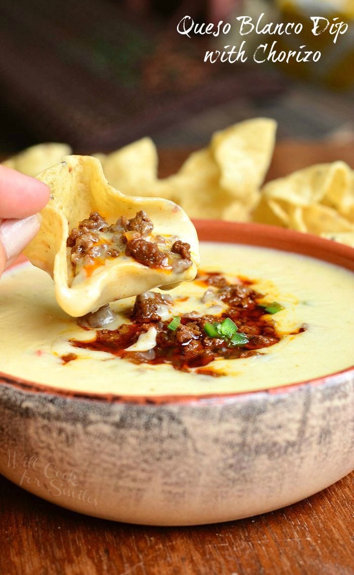 dipping tortilla chip into Queso Blanco Dip with Chorizo in a tan and orange bowl on a wood table 