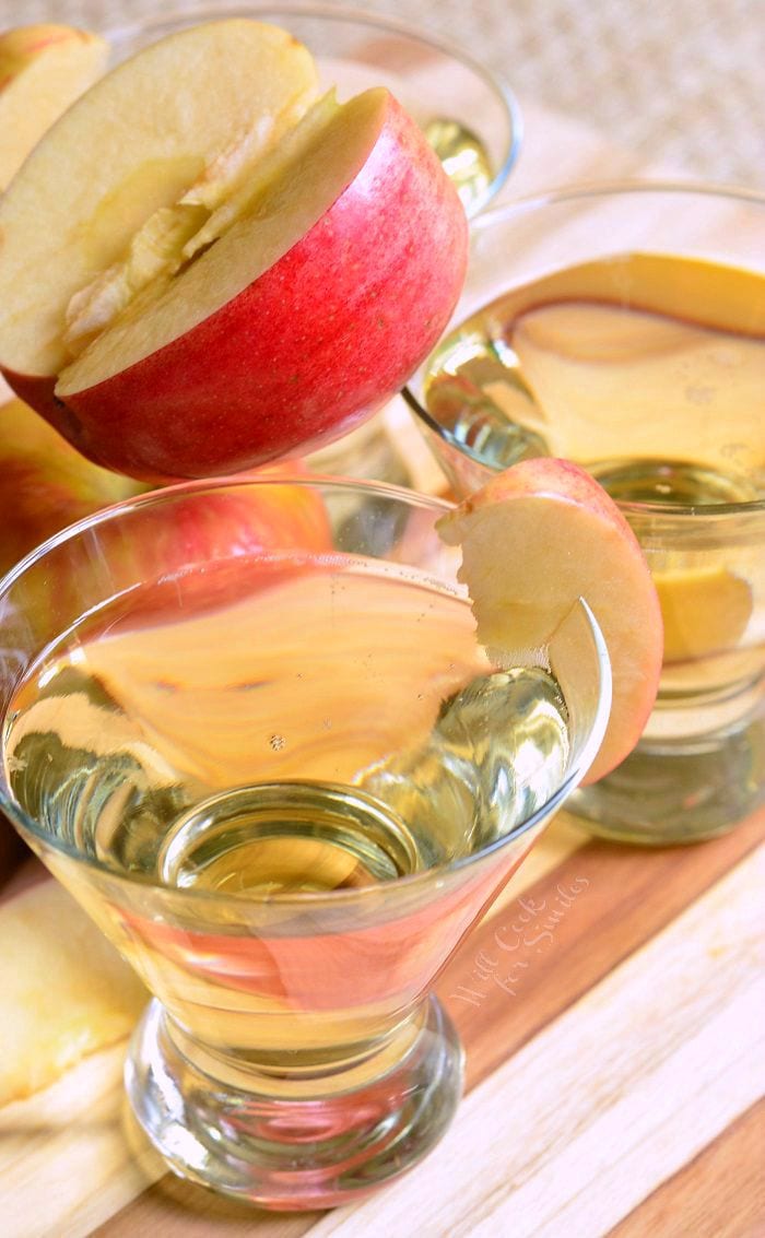 Top view of {Skinny} Cinnamon Apple Champagne Martinis in short martini glasses with a sliced apple placed on the rim of each glass. On the table, there is an apple and apple slice. Balancing on the rims of the glasses and other apple, is a cut red apple.