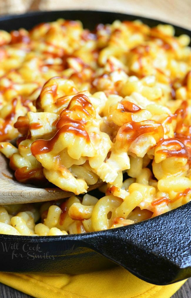 A wooden spoon is dipped into the skillet of Smokehouse Mac and Cheese. There are noodles, chicken, bacon and lots of cheese. The top of the Mac and Cheese is drizzled with BBQ sauce.