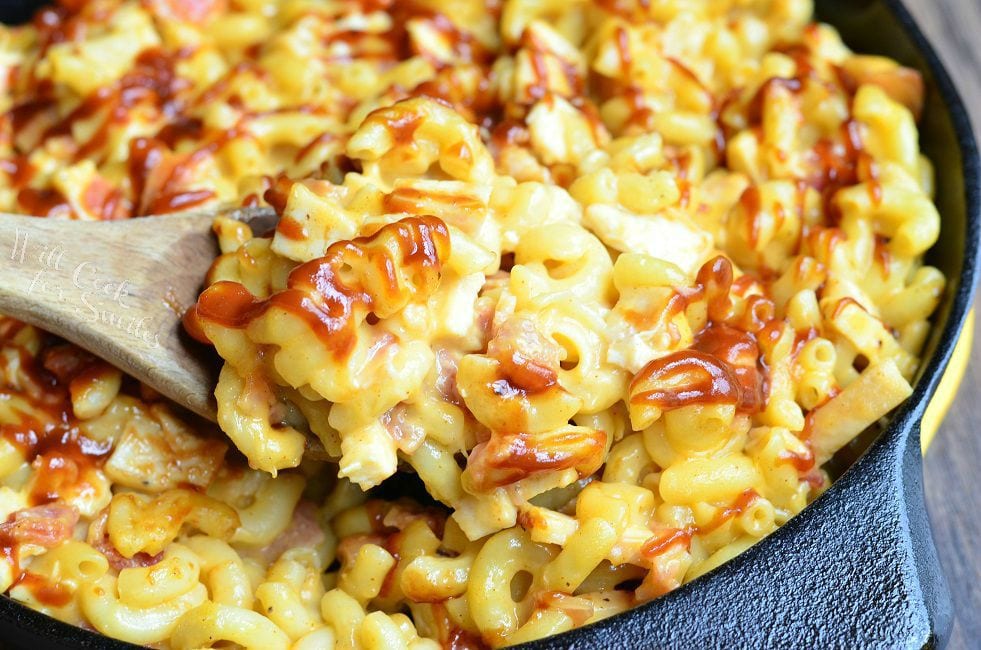 A wooden spoon is dipped into the skillet of Smokehouse Mac and Cheese. There are noodles, chicken, bacon and lots of cheese. The top of the Mac and Cheese is drizzled with BBQ sauce.