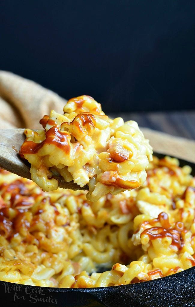 A wooden spoon lifts some of the Smokehouse Mac and Cheese from the black skillet. There are noodles, chicken, bacon and lots of cheese. The top of the Mac and Cheese is drizzled with BBQ sauce.