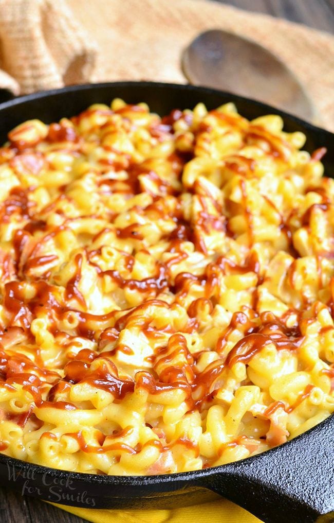 Presented is a black skillet full of Smokehouse Mac and Cheese. There are noodles, chicken, bacon and lots of cheese. The top of the Mac and Cheese is drizzled with BBQ sauce.