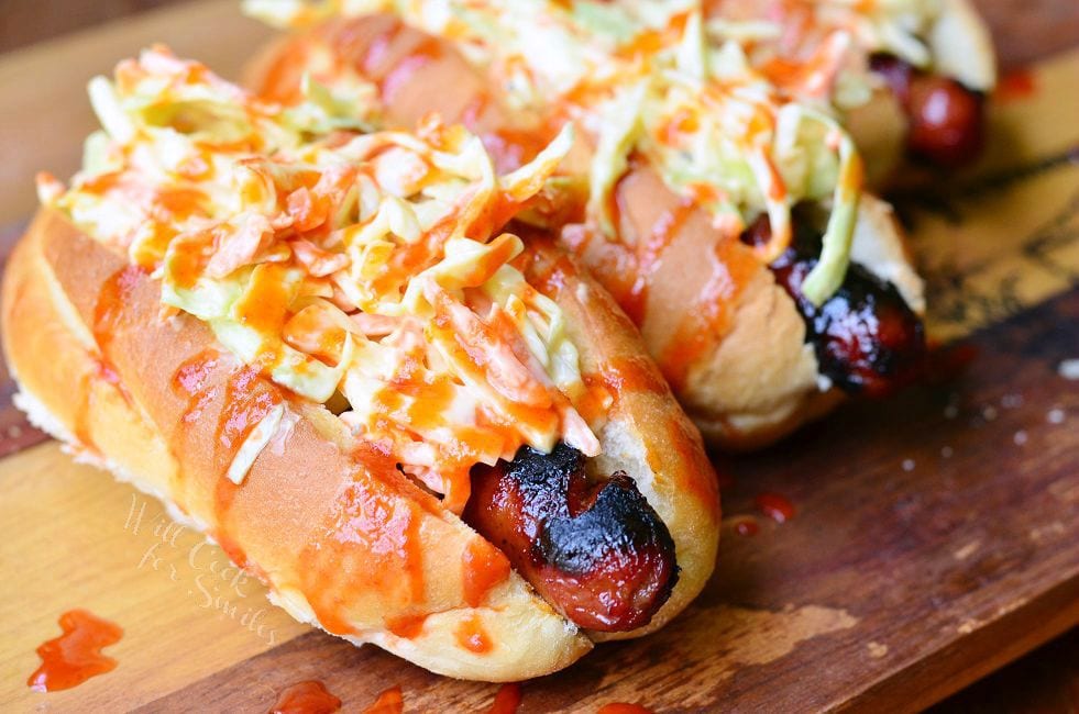 Sriracha Honey Hot Dogs lay side by side on a wood board. Each hot dog bun is packed with a grilled hot dog, coleslaw and then drizzled with the red sriracha honey sauce.