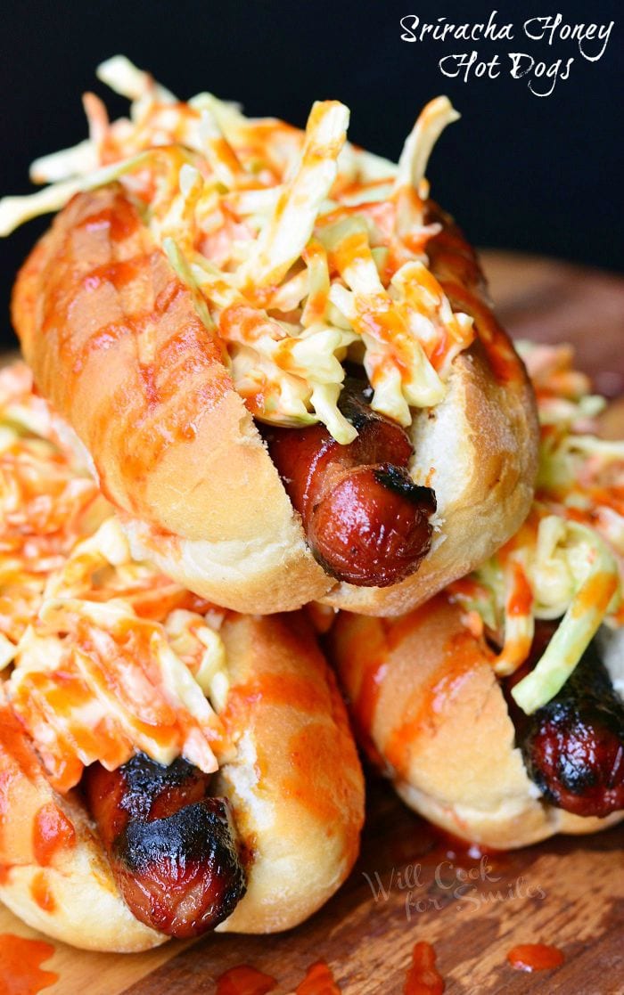 Sriracha Honey Hot Dogs are stacked on each other. Each hot dog bun is packed with a grilled hot dog, coleslaw and then drizzled with the red sriracha honey sauce.