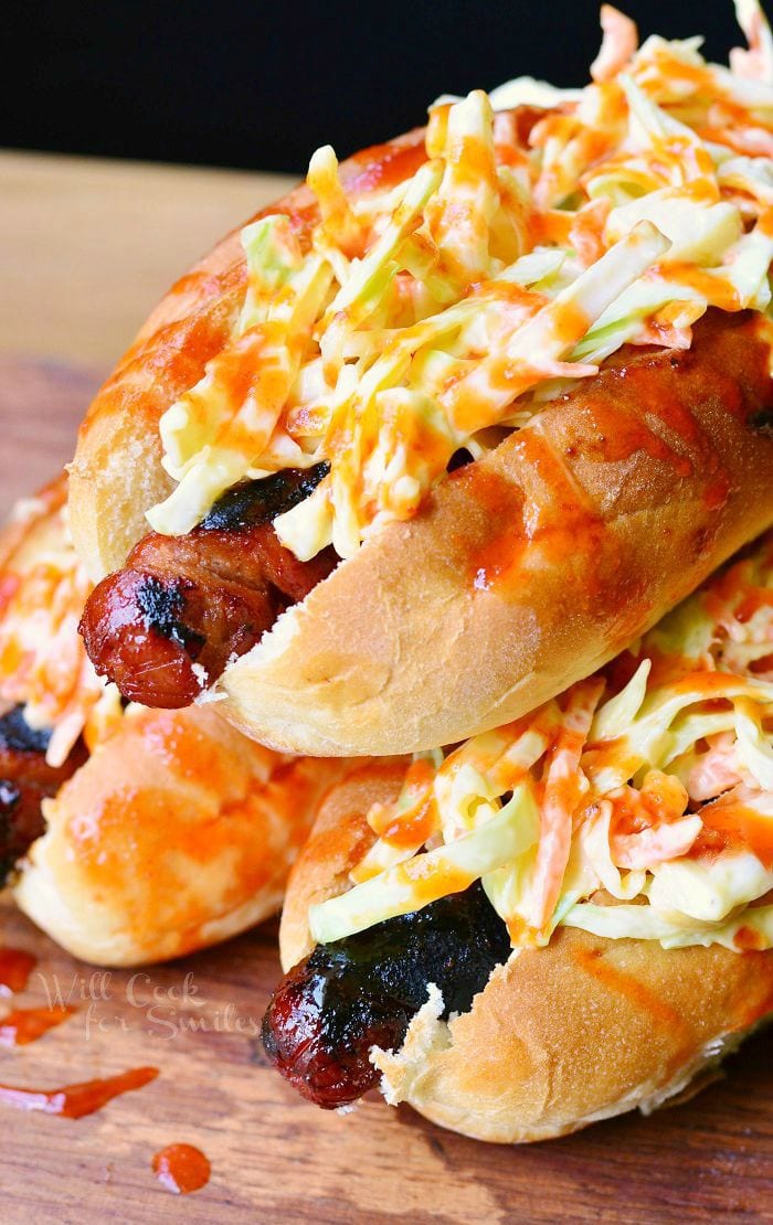 Sriracha Honey Hot Dogs are stacked on each other. Each hot dog bun is packed with a grilled hot dog, coleslaw and then drizzled with the red sriracha honey sauce.