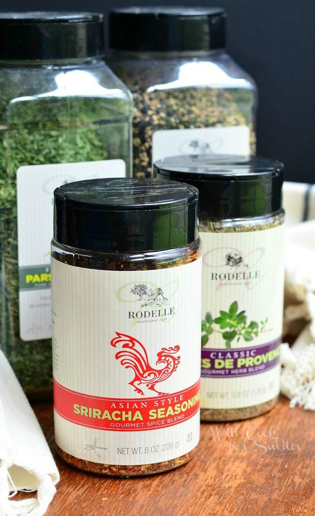 Sriracha Seasoning from Rodelle is in a spice container. The Rodelle label is white with a red tribal dragon. Below the dragon, is a red horizontal stripe saying "Asian Style Sriracha Seasoning". Other Rodelle spice containers are in the background.