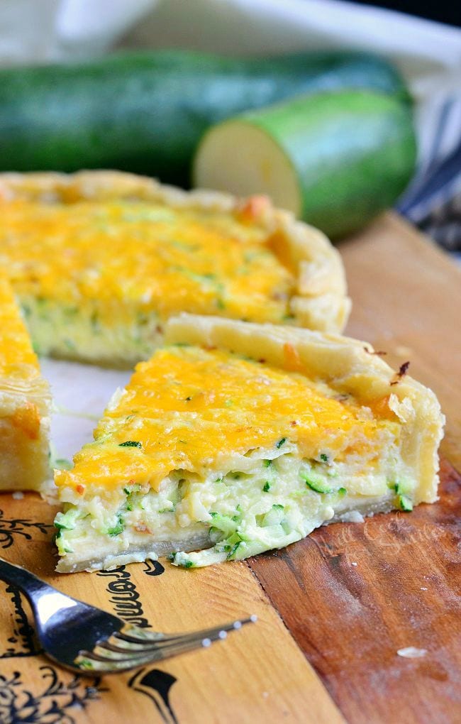 One large piece of Sriracha Zucchini and Cheese Quiche has been cut and moved away from the whole Quiche. The single piece of Quiche is in front at an angle to show the inside of the Quiche. There is also a zucchini in the background.