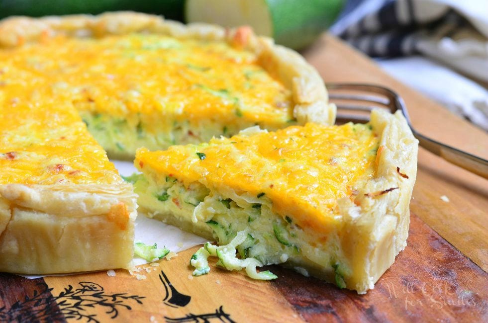 One large piece of Sriracha Zucchini and Cheese Quiche has been cut and moved slightly away from the whole Quiche. This is more of a side view of the Quiche. A fork is laying to the right side of the Quiche.