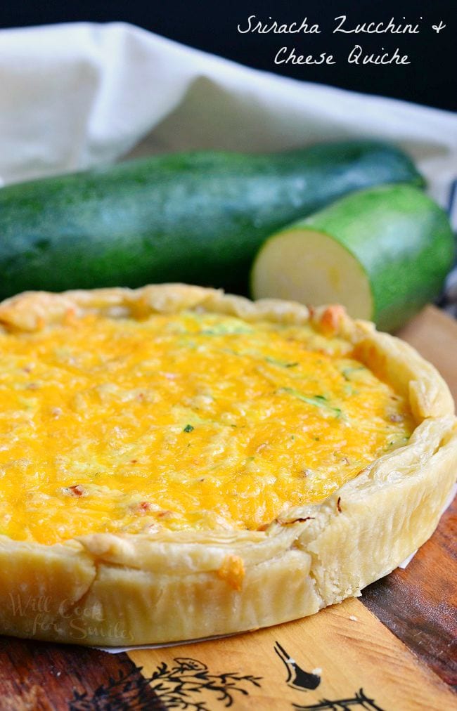 A whole Sriracha Zucchini and Cheese Quiche  is laying on a wood board. The outside crust appears flaky and the top is covered with cheese. There are zucchinis in the background of this photo..