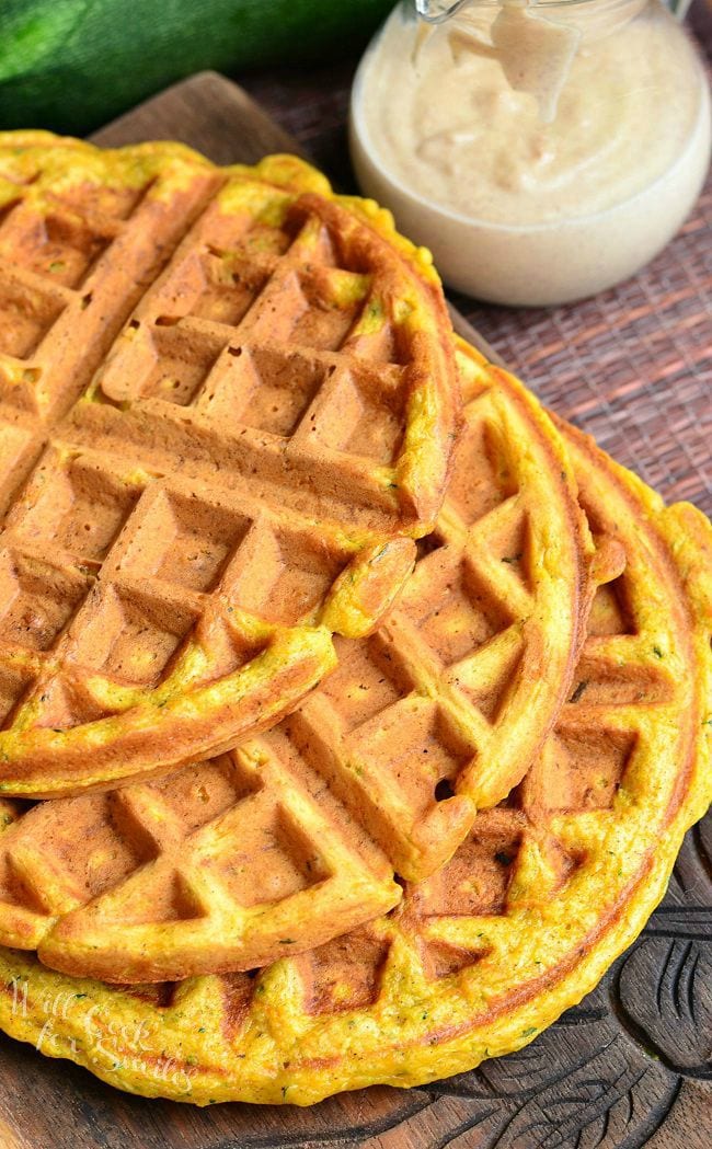 Top view of three Zucchini Carrot Cake Waffles which are laid on a wooden board. The waffles are overlapping each other. Off to the back, right side is a glass container. This container has cinnamon cream cheese sauce in it.