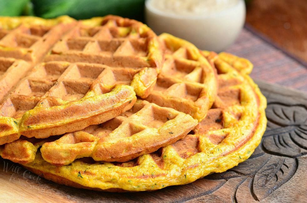 Three Zucchini Carrot Cake Waffles are laid on a wooden board. They are laid in a way that they are overlapping each other.