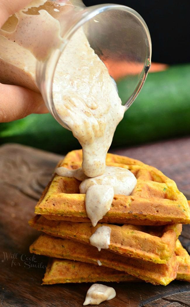 Four pieces of Zucchini Carrot Cake Waffles are stacked up. A glass container is being tilted over the waffles. A cinnamon cream cheese sauce is coming out of the container onto the top of the waffles and dripping down the side.