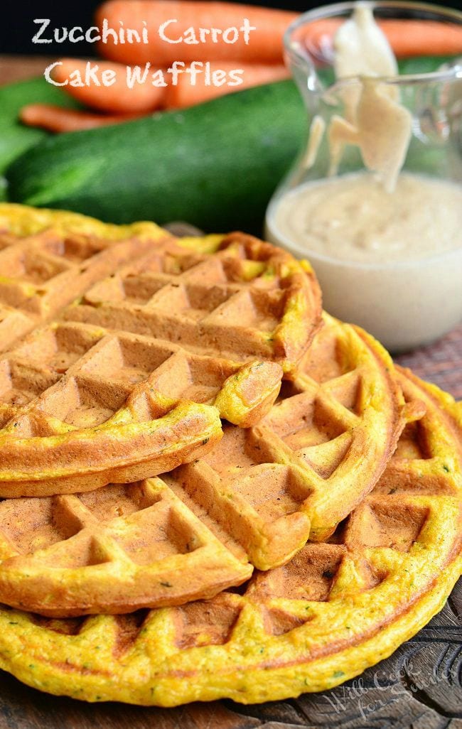 Several Zucchini Carrot Cake Waffles are laid out, overlapping. Behind the waffles is a glass container that contains cinnamon cream cheese sauce.