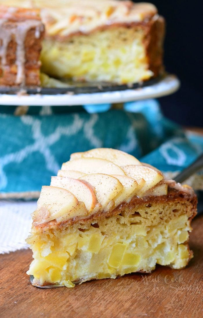 a slice of Cinnamon Glazed Apple Cake with thinly sliced apples on top and a cinnamon glaze on top on a cutting  table with the rest of the cake on a white cake stand 