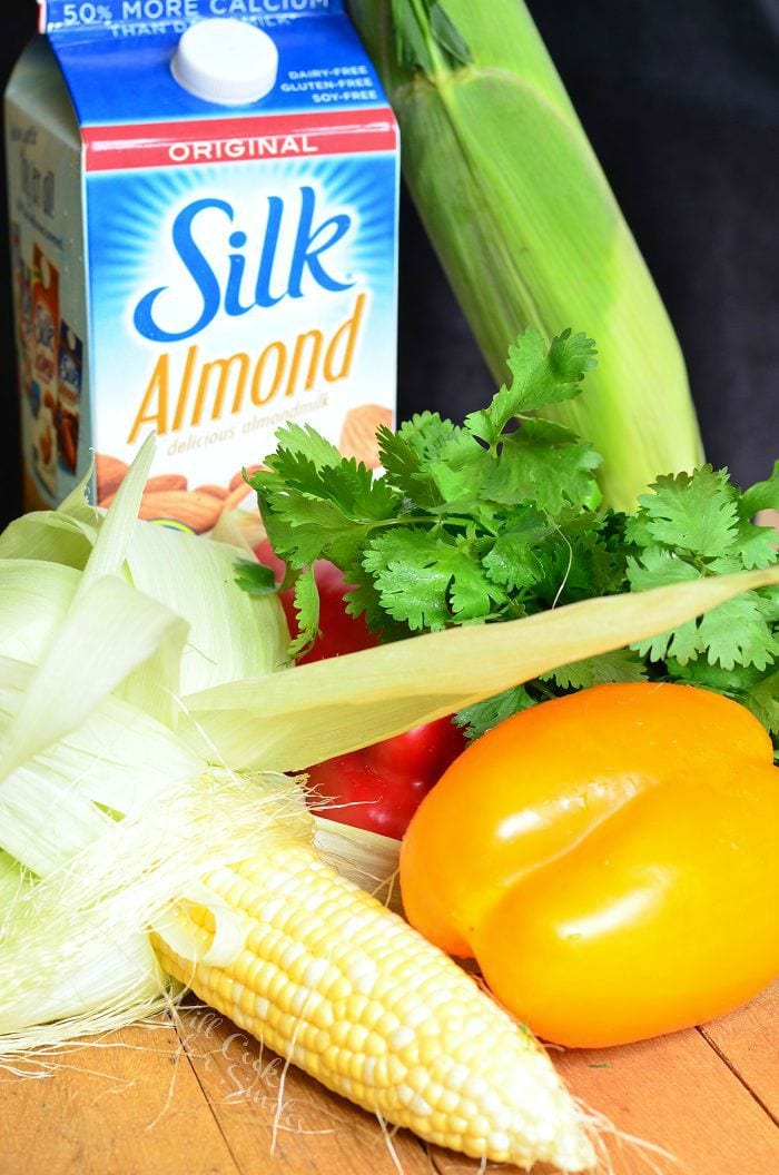 ingredients for dip. Corn with the husk pulled back a yellow and red bell pepper, cilantro, carton of silk almond milk, and a corn on the cob still in the husk all on a table 