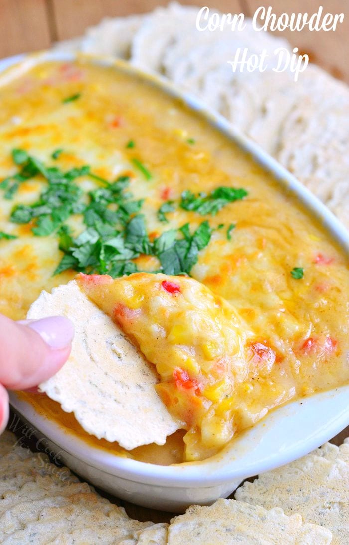 dipping a cracker into Corn Chowder Hot Dip with cilantro on top as a garnish in a white baking dish 