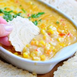 corn chowder hot dip in a white oval dish with crackers around the dish while all sit on a wooden table and a hand dips one cracker into dip viewed close up